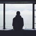 Silhouette of a man looking out of a window at a grey sky , with mental health issues.